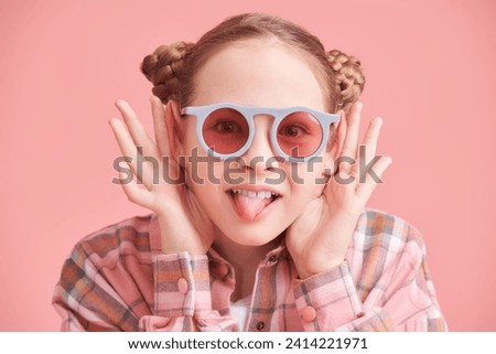 Funny emotional teenage girl in modern clothes and round sunglasses is showing her tounge and teasing. Children, emotions. Pink studio background with copy space.