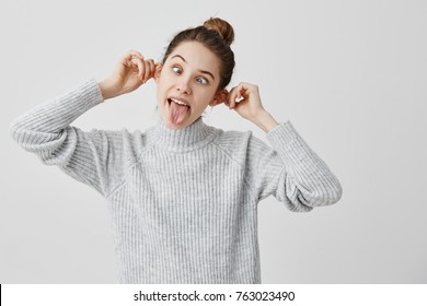 Funny emotions of hilarious female touching her ears and sticking tongue out. Caucasian woman with brown hair in bun pulling face grimacing in studio. Joy, amusement concept 
