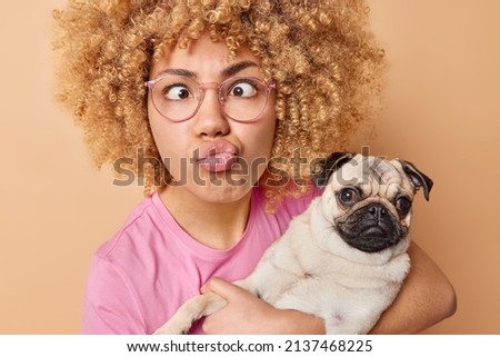Funny European woman with curly bushy hair makes grimace pouts lips and crosses eyes carries pug dog being pet owner wears spectacles pink t shirt isolated over beige background. Animal lover
