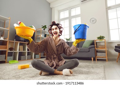 Funny excited woman in hair curlers and clay face mask cleaning house and having fun. Happy energetic young housewife holding plastic bucket and tub sitting on floor at home after doing all housework