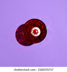 Funny Eye Ball In Red Glass, Creative Horror Inspired Layout On Pastel Purple Background. Halloween Party Idea. 