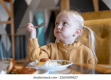 Funny fair-haired girl with two ponytails sits at table holding fork and has breakfast. Mouth full of food. Serious little girl eats