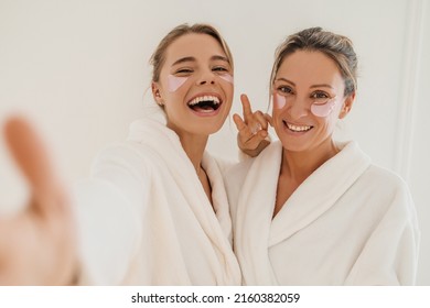 Funny fair-skinned women in white coats with wide smile look at camera indoors. Blondes wear eye patches for dark circles. Beauty needs care concept