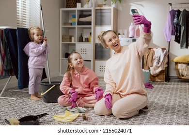 Funny Family Having Fun While Doing Housework And Cleaning. Smiling Woman Taking Selfie With Daughters. Happy Girl Uses Smartphone To Talk Video Shows Kids Helping Her Clean Bathroom.