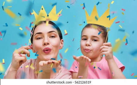 Funny Family On A Background Of Bright Blue Wall. Mother And Her Daughter Girl Blow Confetti. Mom And Child Are Holding Paper Crowns On Stick.