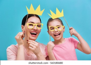 Funny Family On A Background Of Bright Blue Wall. Mother And Her Daughter Girl With A Paper Accessories. Mom And Child Are Holding Crowns Glasses And On Stick.