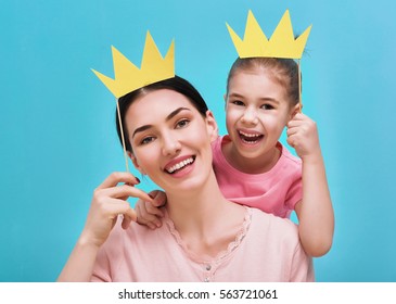 Funny Family On A Background Of Bright Blue Wall. Mother And Her Daughter Girl With A Paper Accessories. Mom And Child Are Holding Crown On Stick.