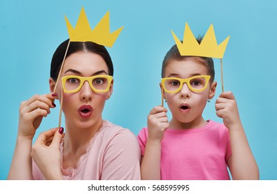 Funny Family On A Background Of Bright Blue Wall. Mother And Her Daughter Girl With A Paper Accessories. Mom And Child Are Holding Crowns Glasses And On Stick.