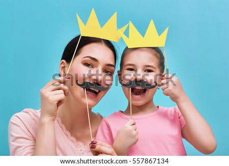 Funny family on a background of bright blue wall. Mother and her daughter girl with a paper accessories. Mom and child are holding paper crown and mustache on sticks.
