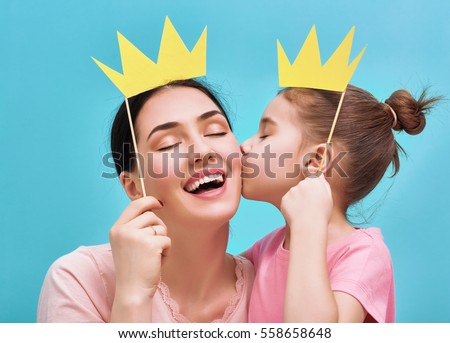Funny family on a background of bright blue wall. Mother and her daughter girl with a paper accessories. Mom and child are holding paper crown on stick.