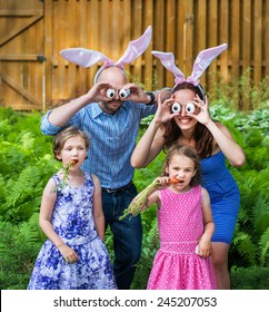 Funny Family Portrait On Easter Of Mother And Father In Bunny Ears And Silly Eyes Made From Eggs As Their Children Pose Eating Carrots Outside In A Garden During The Spring Season. Part Of A Series.