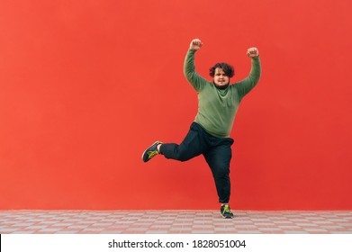 Funny Fat Guy Dancing On A Red Wall And Listening To Music On Headphones. Cheerful Overweight Man And Casual Clothes Shows A Dance Performance.