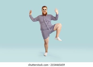 Funny fat guy enjoying his happy summer holiday. Full body length portrait of cheerful goofy chubby bearded man wearing striped retro swimsuit and sunglasses dancing against blue studio background