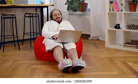 Funny Fat Guy Wearing Bathrobe And Unicorn Slippers Sitting In Beanbag And Using Laptop. Portrait Of Overweight Man Resting In Living Room At Home And Working On Computer