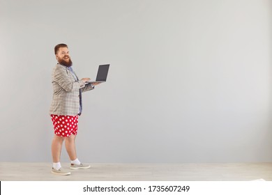 Funny fat man in a blue shirt and red shorts standing working online using a laptop on a gray background.