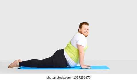 A Funny Fat Man Does Yoga Exercises On A Mat On A Background.