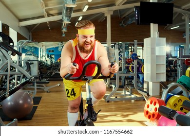 A Funny Fat Man  Doing Exercises On An Exercise Bike In A Sports