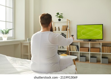 Funny fat man in pyjamas sitting on bed at home and watching football on television, back view. Lazy chubby ginger guy wakes up in morning and switches soccer on sports TV channel with remote control