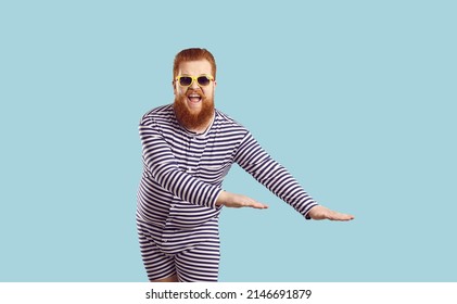 Funny fat man in retro swimsuit enjoying his summer holiday. Studio portrait of happy goofy chubby bearded guy wearing striped swimming suit and sunglasses dancing isolated on blue color background