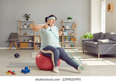 Funny fat woman doing home workouts. Overweight lady doing sports exercise. Chubby woman in T shirt and leggings sitting on fit ball in living room and doing difficult exercise with resistance band - Shutterstock ID 2299035995