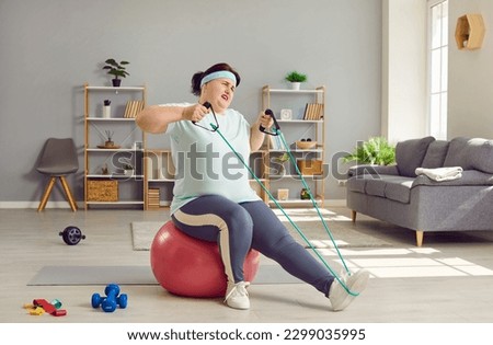 Funny fat woman doing home workouts. Overweight lady doing sports exercise. Chubby woman in T shirt and leggings sitting on fit ball in living room and doing difficult exercise with resistance band