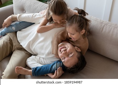 Funny Father Pretends To Be Tired Or Asleep While Two Pretty Daughters Want To Play. Happy Family Lying On Couch At Home Enjoy Playtime Active Weekends Together, Having Fun, Games With Kids Concept