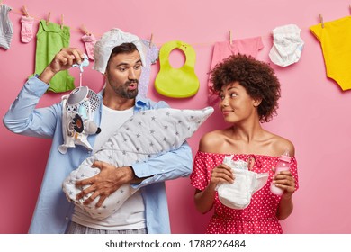 Funny Father Tries To Sooth Newborn, Shows Nipple And Talks To Baby, Wears Diaper On Head, Happy Cheerful Mother With Nappy And Feeding Bottle In Hands. Glad Young Parents Watch Their Baby Sleeping.