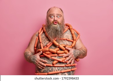 Funny fatso male keeps lips rounded, poses with naked body wrapped by sausages, has health problems connected with health and unhealthy nutrition, likes meat, stands indoor. Gluttony concept