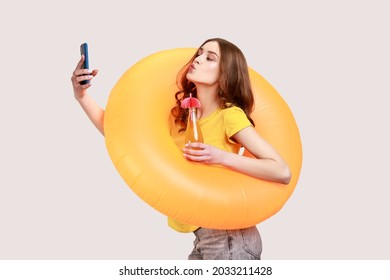 Funny Female With Brown Wavy Hair In Yellow T-shirt Holding Orange Rubber Ring And Making Selfie At Phone Makes Kiss Gesture, Having Rest On Resort. Indoor Studio Shot Isolated On Gray Background.