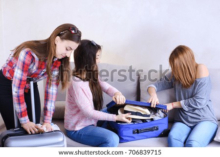 Funny female friends together collect large gray and blue suitcases, add up all necessary things for class holiday in another country on sea. Young women fool around and laugh, measure glasses and
