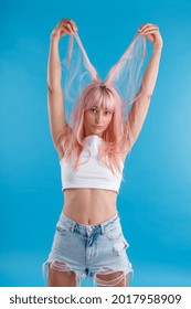 Funny female model in casual wear playing with pink hair, holding strands of it up over her head, standing isolated over blue studio background. Beauty, hair care concept