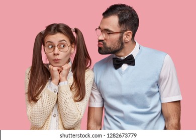Funny Female Nerd With Two Pony Tails, Wears Big Spectacles, Going To Recieve Kiss From Boyfriend, Has First Date, Feels Awkward, Isolated Over Pink Background. Bearded Guy In Elegant Clothes Flirts