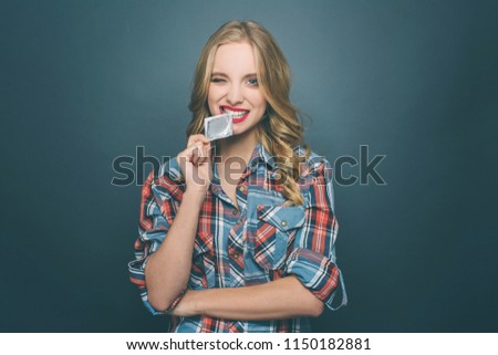 Funny firl is biting a piece of condom's package. She is looking straight forward. Isolated on blue background