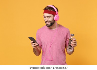 Funny Fitness Sporty Guy 20s Sportsman In Headband T-shirt In Home Gym Isolated On Yellow Background. Workout Sport Motivation Concept. Hold Water Bottle, Listen Music With Headphones, Mobile Phone