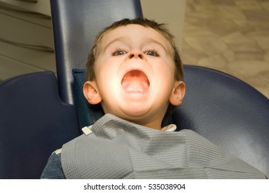 Funny Five Y.o. Boy With Wide Opened Mouth Is Sitting In The Dentist Chair Under The Medical Lamp Light  Ready To Treat His Teeth