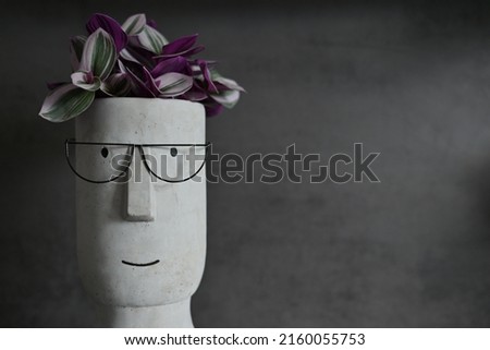 Funny flowerpot made of concrete with smiling face wearing glasses containing a flower in front of a grey or charcoal backdrop.