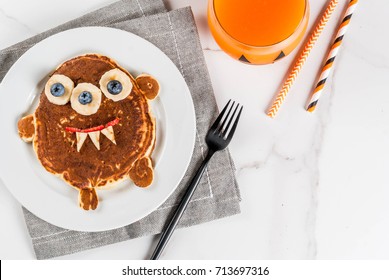 Funny Food For Halloween. Kids Breakfast Pancake Decorated Like Creepy Monster, With Banana, Berries, With Pumpkin Smoothie Juice, White Table Copy Space Top View