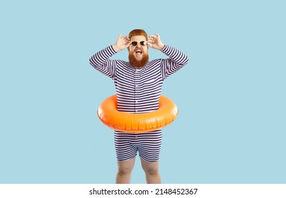 Funny funky cheerful man enjoying summer vacation isolated pastel turquoise background. Joyfully excited red-bearded guy in striped leotard and with inflatable swimming circle adjusts his sunglasses
