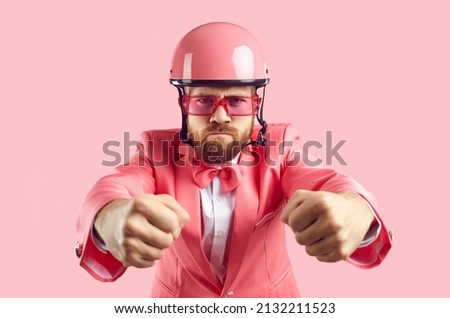 Funny furious tough driver holding pretend steering wheel. Studio shot of man with ginger beard and angry face, wearing pink helmet, funky modern suit, bow tie and sunglasses driving his invisible car