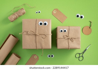 Funny gift boxes with eyes and wrapping materials. Jute rope, wrapping paper roll, scissors and gift tags on bright green background.
