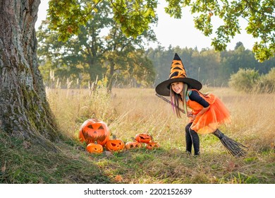 A Funny Girl In A Big Hat And An Orange Dress In The Image Of A Witch Is Sitting On A Broomstick, Laughing Merrily. Decorative Carved And Painted Sinister Pumpkins For Halloween