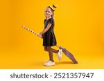 Funny girl with broom dressed like witch on yellow background. Halloween celebration