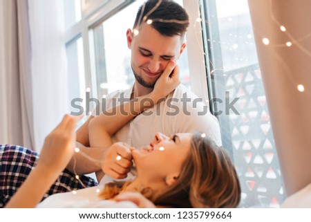 Funny girl in cute pajama touching her boyfriend's face. Cheerful brunette male model looking at blonde woman with love.