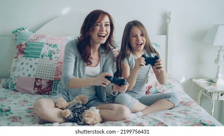 Funny Girl With Happy Young Mother Have Fun While Playing Computer Console Games On TV Sitting On Bed At Home In The Morning In Cozy Bedroom. Daughter Embracing Mom After Winning