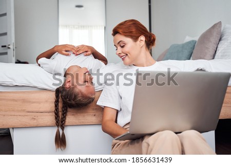 Funny girl having fun with her mother at the bedroom while woman sitting and working with laptop. Parenthood concept