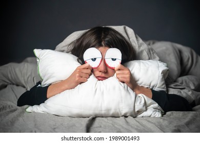 A Funny Girl Hugs A Pillow, Holds Drawn Creative Tired Red Eyes In Her Hands, Covered With A Blanket. A Woman Is Worried About Insomnia, Poor Sleep, Stress.