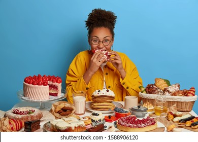 Funny glutton woman bites cakes with big appetite, cant stop eating sweet desserts, being in mood for enjoying sugary freshly baked bakery, surrounded by sweet treasures, has yummy snack, feels hunger