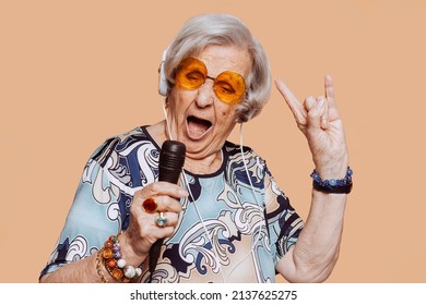 Funny Grandmother Portrait Doing Horns Sign Gesture With Hand While Singing Karaoke Using Microphone And Headphones At Studio. Senior Old Woman Dressing Elegant For A Special Event. Rockstar Granny.