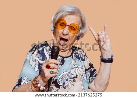 Funny grandmother portrait doing horns sign gesture with hand while singing karaoke using microphone and headphones at studio. Senior old woman dressing elegant for a special event. Rockstar granny.