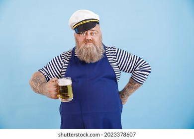 Funny grimacing plus size man in sailor suit with apron and white hat holds glass of foamy beer on light blue background in studio
